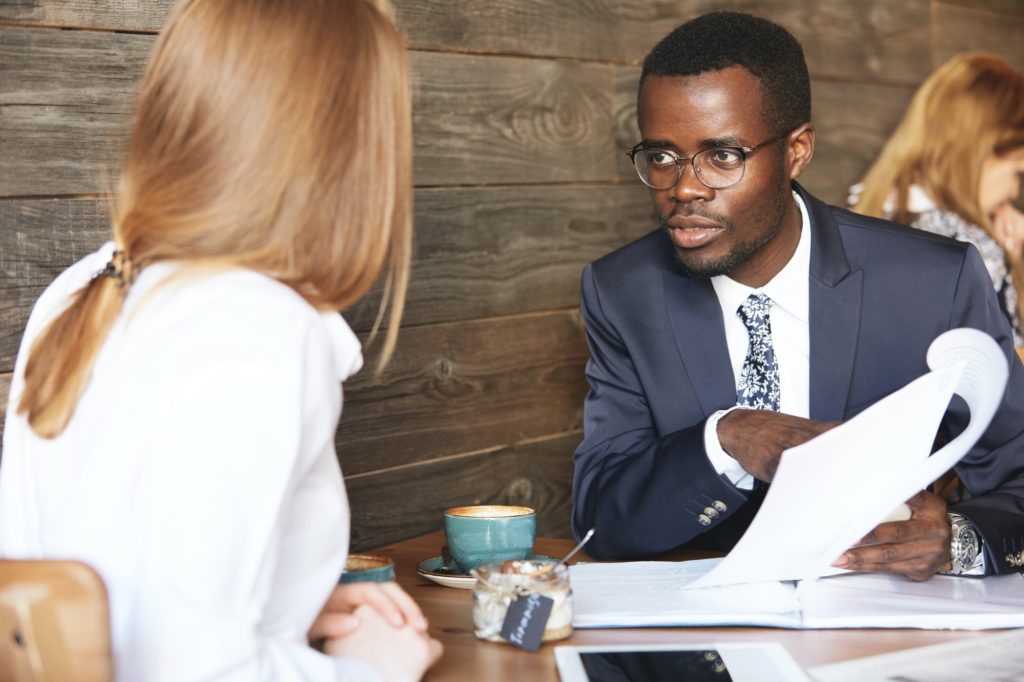 African boss conducting an interview with redhead Caucasian woman, holding job application and liste