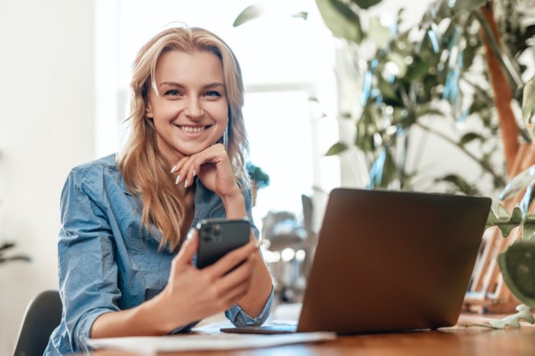 Glad blond haired woman sitting at table with laptop and mobilephone