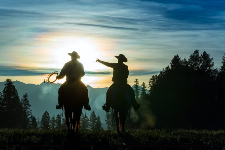 Two cowboys riding across grassland with mountains in background, early morning